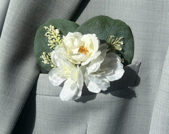 May include: A white floral boutonniere with green leaves and sprigs, set against a light gray suit jacket. The boutonniere features a large, multi-petaled flower with a yellow center.