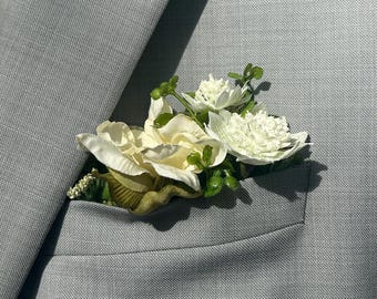 May include: A light gray suit jacket with a pocket holding a floral boutonniere. The boutonniere features white and cream-colored flowers with green accents. The jacket's fabric has a subtle texture.