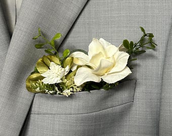 May include: A white and green floral boutonniere in the pocket of a light gray suit jacket. The boutonniere features a large white flower, smaller white flowers, and green foliage.