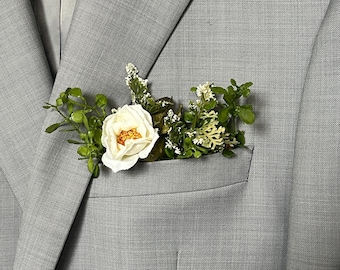 May include: A white and orange flower boutonniere with green leaves and small white flowers in the pocket of a light gray suit jacket. The floral arrangement adds a touch of elegance.
