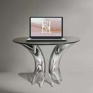 May include: A silver, abstract, modern table with a glass top. A laptop computer is on the table with a screen displaying a light brown and white background with the letters "EI" and a small floral design.
