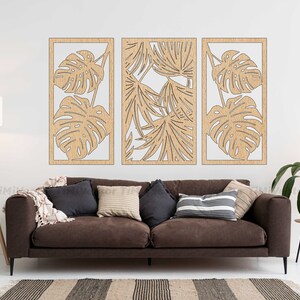 May include: Three wooden wall panels with a cutout design of tropical leaves. The panels are arranged in a row on a white wall, above a brown sofa with pillows.