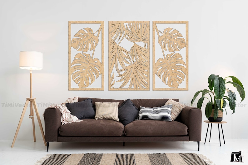 May include: Three wooden wall panels with a cutout design of tropical leaves. The panels are arranged in a row on a white wall, above a brown sofa with pillows.