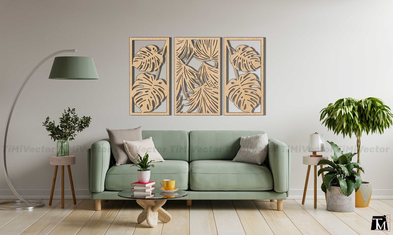 May include: Three wooden wall panels with a cutout design of tropical leaves. The panels are arranged horizontally and are mounted on a white wall. A green sofa with pillows is in the foreground.
