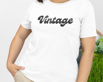 May include: A white short-sleeved crew neck t-shirt featuring the word Vintage printed in a black, distressed, cursive font across the chest. The shirt has a standard fit with a straight hemline and is made of a solid white fabric.