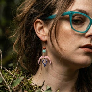 May include: Gold drop earrings with a celestial design. The earrings feature a pink crescent moon, a blue female symbol, and three beads in brown and turquoise. The earrings are designed with a drop style.