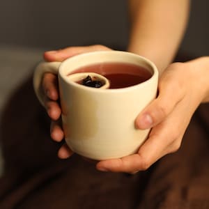 Puede incluir: Una persona sostiene una taza de cerámica blanca llena de una bebida roja. La taza tiene un diseño único con una pequeña abertura redonda en el lateral.