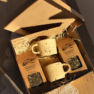 Könnte beinhalten: Eine Geschenkbox gefüllt mit zwei gesprenkelten, beigefarbenen Keramikbechern und zwei Packungen Kräutertee mit der Aufschrift "Чай трав'яний "Вишиванка". Die Box ist mit zerkleinertem Papier ausgelegt.