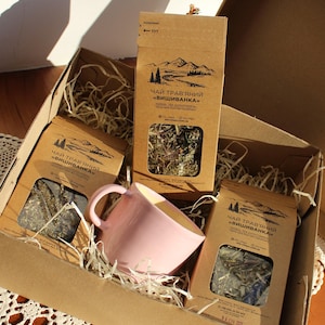 Könnte beinhalten: Eine Geschenkbox aus Pappe mit drei Kräuterteepäckchen und einer rosa Keramik-Tasse. Die Teepäckchen sind braun mit einem Berg- und Baumdesign und dem Text "Herbal Tea". Die Tasse ist hellrosa. Die Box ist mit Stroh gefüllt.