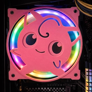 May include: A pink computer fan featuring a cartoon character design. The fan has a circular frame with a smiling face and large eyes. The fan blades are illuminated with a rainbow of colours, creating a vibrant effect. The fan is mounted inside a computer case.
