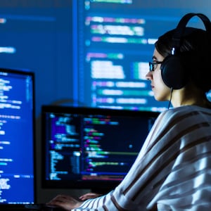 Può includere: Una donna con cuffie e occhiali è seduta davanti a uno schermo di computer che mostra codice. Lo schermo è blu e il codice è bianco. La donna sta digitando su una tastiera.