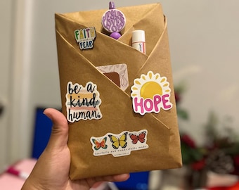 May include: A brown paper-wrapped gift decorated with stickers. Stickers include the phrases "Faith over Fear", "be a kind human", and "HOPE" with a sun graphic. Also, butterfly stickers and a lip balm are visible.