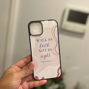 Puede incluir: Funda de teléfono negra con el texto "WALK BY faith, NOT BY sight" y el versículo bíblico 2 Corintios 5:7. La funda tiene un fondo abstracto rosa y blanco. La funda del teléfono está en una mano.