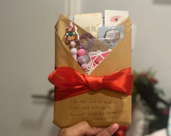 May include: A gift wrapped in brown paper, secured with a red satin bow. The package contains small items like cards and beaded accessories. Text on the package reads: "For the word of God is right and true; He is faithful in all all we does." Psalms 33:4.