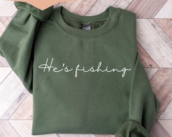 Könnte beinhalten: Ein grüner Pullover mit dem Schriftzug "He's fishing" in weißer Schrift. Der Pullover ist gefaltet und auf einer Holzoberfläche abgelegt.