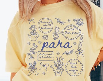 Może przedstawiać: Bladożółty t-shirt z ciemnoniebieskim nadrukiem graficznym przedstawiającym różne ilustracje kwiatowe, kokardy i konewki. Tekst zawiera "learning with kindness", "watch them grow", "para", "planting seeds of knowledge" oraz "teach love inspire".