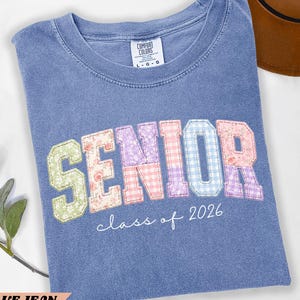 May include: Blue jean-colored t-shirt with the word "SENIOR" in colorful patchwork letters. The shirt also features "class of 2026" in a cursive font. The shirt is folded and has a Comfort Colors tag.