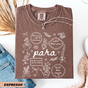 May include: A brown t-shirt featuring white line art illustrations of flowers, a teapot, a watering can, and bows. The text reads "learning with kindness," "watch them grow," "para," "planting seeds of knowledge," and "teach love inspire."