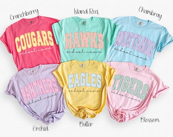 Könnte beinhalten: Sechs kurzärmelige T-Shirts in Pink, Mint, Hellblau, Lavendel, Gelb und Hellrosa. Jedes T-Shirt zeigt einen Schulnamen in einer gestreiften Schriftart mit dem Text school name in Schreibschrift darunter. Die Farben sind als Crunchberry, Island Reef, Chambray, Orchid, Butter und Blossom beschriftet.