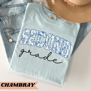 May include: A light blue t-shirt with the words "Second grade" in a blue and white floral print. The shirt is made of a soft, comfortable fabric and is perfect for school or everyday wear.