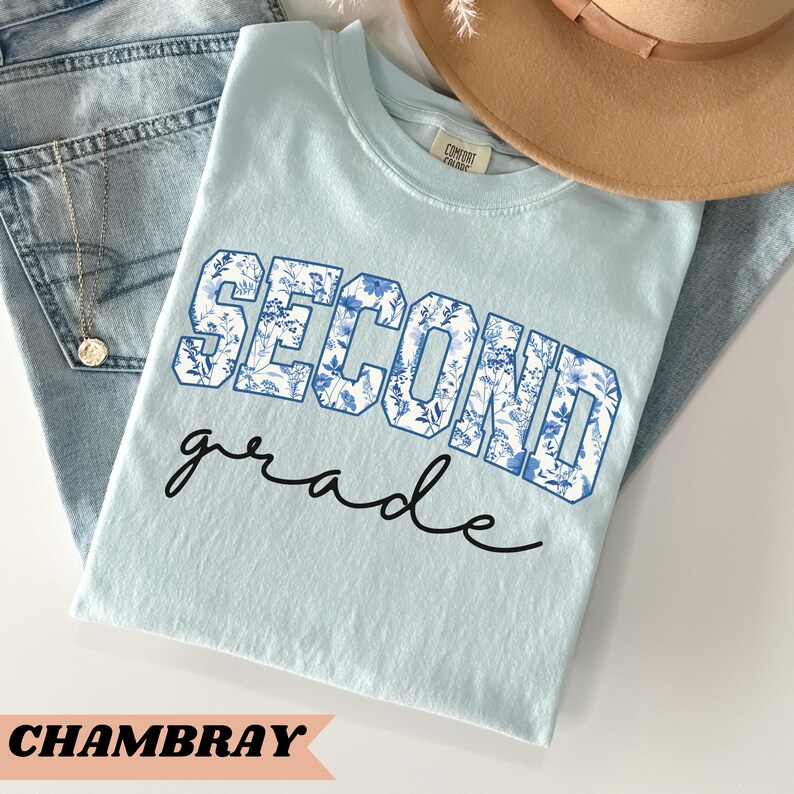 May include: A light blue t-shirt with the words "Second grade" in a blue and white floral print. The shirt is made of a soft, comfortable fabric and is perfect for school or everyday wear.
