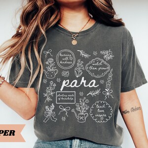 May include: A charcoal gray short-sleeve t-shirt featuring a white line-art graphic with floral elements, watering cans, and text including "learning with kindness," "watch them grow," "para," "planting seeds of knowledge," and "teach love inspire."