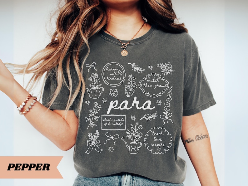 May include: A charcoal gray short-sleeve t-shirt featuring a white line-art graphic with floral elements, watering cans, and text including "learning with kindness," "watch them grow," "para," "planting seeds of knowledge," and "teach love inspire."