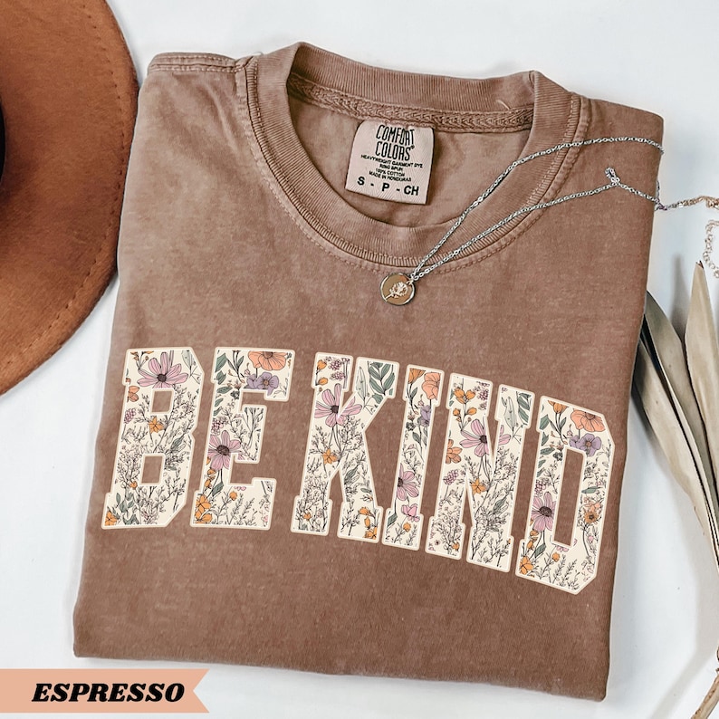May include: Espresso-colored t-shirt with the words "BE KIND" in a floral design. The shirt has a crew neck and short sleeves. A silver necklace with a pendant is draped across the shirt. The shirt is folded on a white surface.