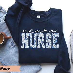 Puede incluir: Sudadera azul marino con un dise&ntilde;o floral blanco y el texto "neuro nurse".