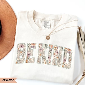 May include: Ivory-colored t-shirt with the words "BE KIND" in a floral design. The shirt has a crew neck and short sleeves. A gold necklace with a pendant rests on the shirt. The shirt is labeled "Comfort Colors".