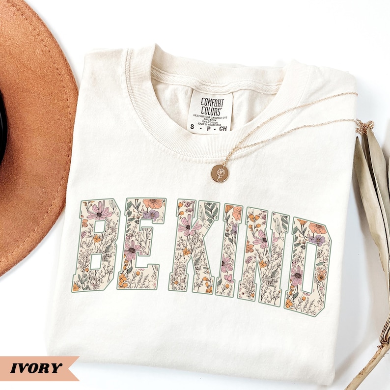 May include: Ivory-colored t-shirt with the words "BE KIND" in a floral design. The shirt has a crew neck and short sleeves. A gold necklace with a pendant rests on the shirt. The shirt is labeled "Comfort Colors".