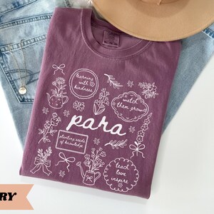 May include: A berry-colored t-shirt featuring white line art illustrations of flowers, a teapot, a watering can, and bows. The text reads "learning with kindness," "watch them grow," "para," "planting seeds of knowledge," and "teach love inspire."