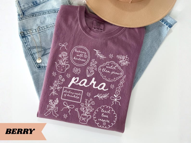 May include: A berry-colored t-shirt featuring white line art illustrations of flowers, a teapot, a watering can, and bows. The text reads "learning with kindness," "watch them grow," "para," "planting seeds of knowledge," and "teach love inspire."