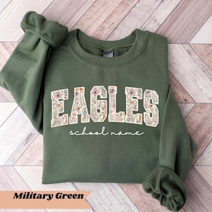 May include: Military green sweatshirt with the word "EAGLES" in a floral design. Below the word "EAGLES" is the text "school name". The sweatshirt is on a light brown wooden surface.
