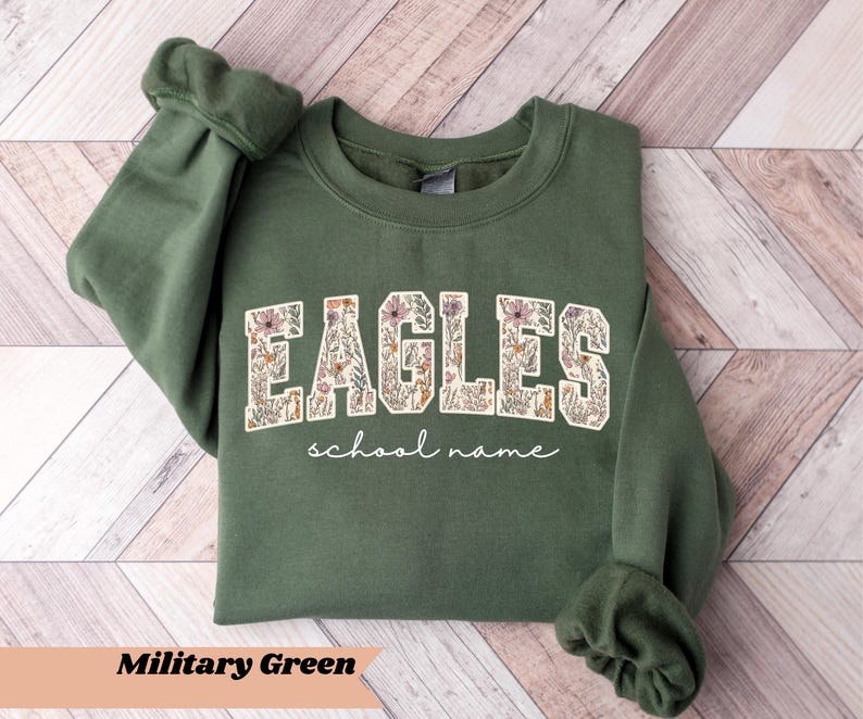 May include: Military green sweatshirt with the word "EAGLES" in a floral design. Below the word "EAGLES" is the text "school name". The sweatshirt is on a light brown wooden surface.