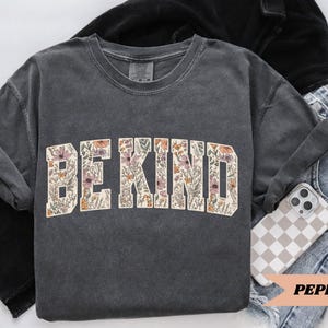 May include: A dark gray sweatshirt with the words "BE KIND" in a floral design. The sweatshirt is folded with a pair of blue jeans and a phone with a checkered case. The word "PEPPER" is printed on a peach-colored banner.