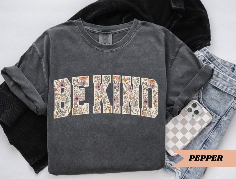 May include: A dark gray sweatshirt with the words "BE KIND" in a floral design. The sweatshirt is folded with a pair of blue jeans and a phone with a checkered case. The word "PEPPER" is printed on a peach-colored banner.
