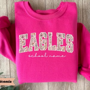 May include: A bright pink sweatshirt with the word "EAGLES" in a floral design across the chest. Below the word "EAGLES" is the phrase "school name" in a cursive font. The sweatshirt has long sleeves and a crew neck.