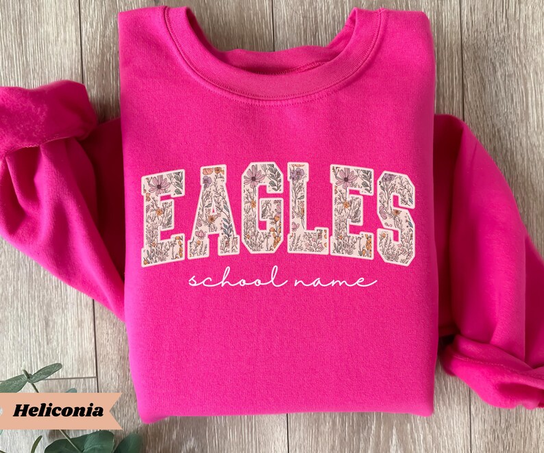 May include: A bright pink sweatshirt with the word "EAGLES" in a floral design across the chest. Below the word "EAGLES" is the phrase "school name" in a cursive font. The sweatshirt has long sleeves and a crew neck.