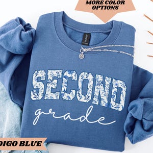 May include: Indigo blue sweatshirt with a white floral design that says "Second grade".