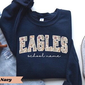 May include: Navy blue sweatshirt with the word "EAGLES" in a floral design. Below the word "EAGLES" is the phrase "school name" in a cursive font. The sweatshirt is folded on top of a pair of blue jeans and a pair of gray sneakers.