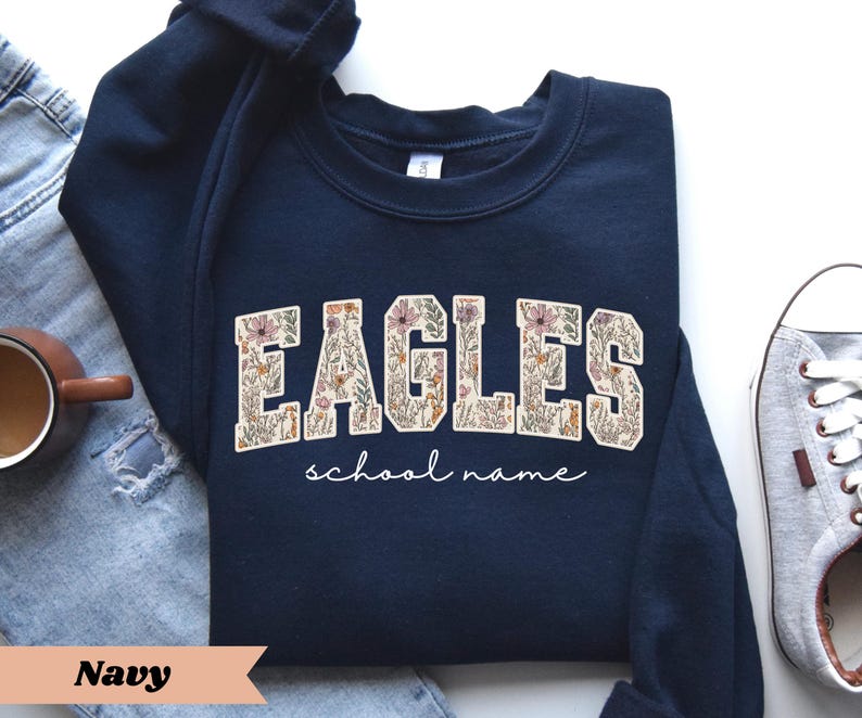 May include: Navy blue sweatshirt with the word "EAGLES" in a floral design. Below the word "EAGLES" is the phrase "school name" in a cursive font. The sweatshirt is folded on top of a pair of blue jeans and a pair of gray sneakers.