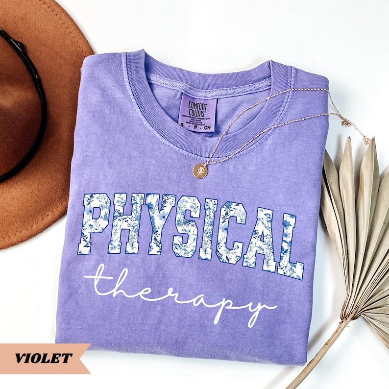 May include: A violet Comfort Colors t-shirt with the words "PHYSICAL" and "therapy" printed on the front. The word "PHYSICAL" is in a blue floral design. A gold necklace and a brown hat are also in the image.