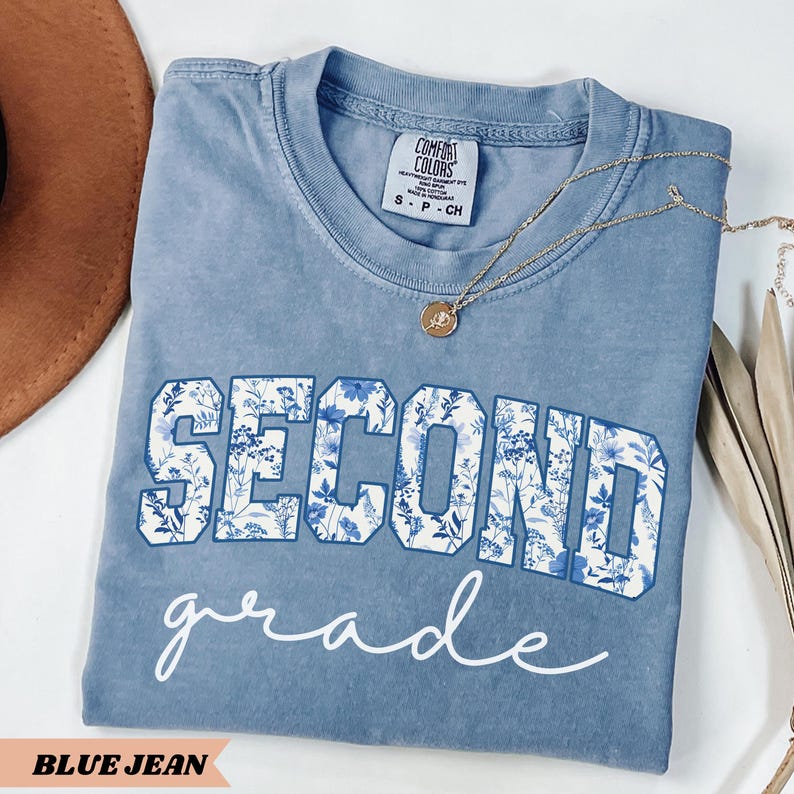 May include: A blue jean t-shirt with the word "SECOND" in white letters with a blue floral pattern. The word "grade" is written in white cursive below the word "SECOND".