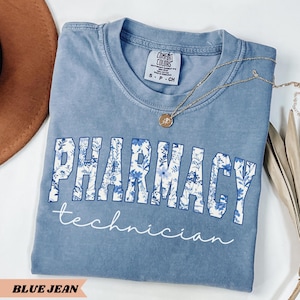 May include: A blue jean colored t-shirt with the words "PHARMACY technician" in white and blue floral print. The shirt has a crew neck and short sleeves. A gold necklace with a pendant is draped on the shirt.