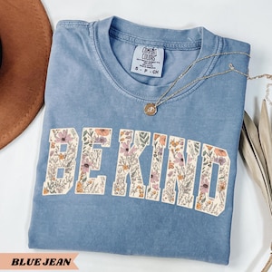 May include: A blue jean colored t-shirt with the words "BE KIND" in a floral design. The shirt has a crew neck and short sleeves. A gold necklace with a pendant is draped on the shirt.