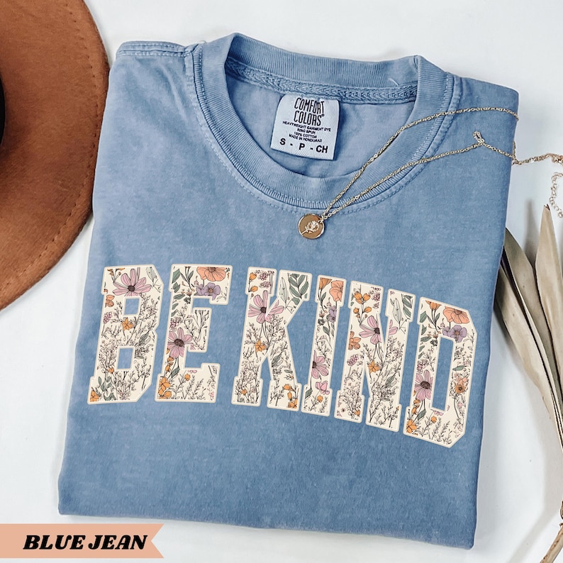 May include: A blue jean colored t-shirt with the words "BE KIND" in a floral design. The shirt has a crew neck and short sleeves. A gold necklace with a pendant is draped on the shirt.