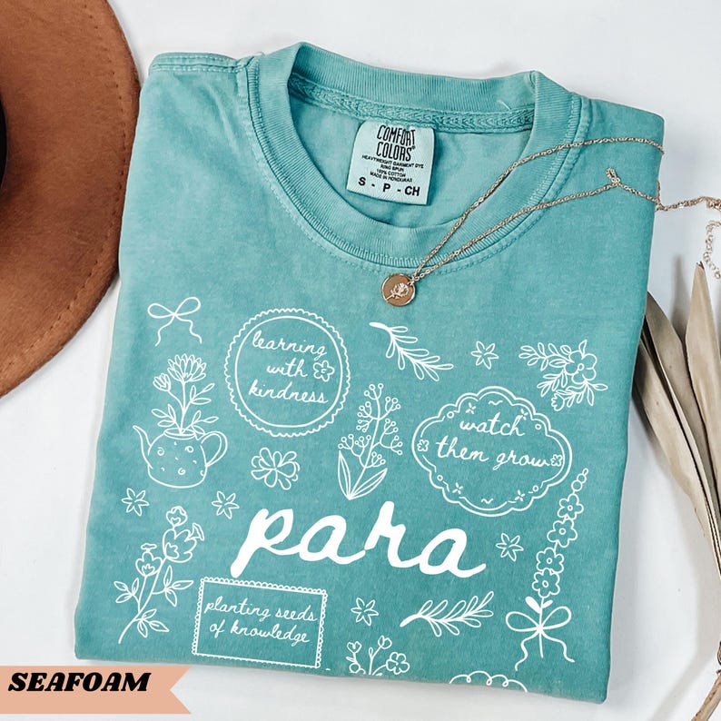 May include: A teal short-sleeved t-shirt featuring white line art of flowers, a teapot, and text that reads learning with kindness, watch them grow, para, and planting seeds of knowledge. A gold-toned necklace with a small circular pendant rests on top.