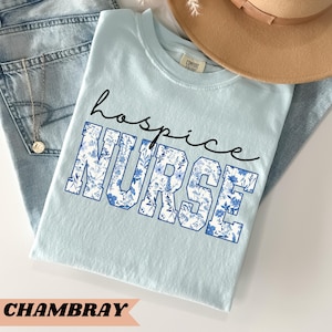 May include: Light blue t-shirt with the text "hospice NURSE" in white letters with a blue floral pattern. The shirt is made of a soft, comfortable fabric.