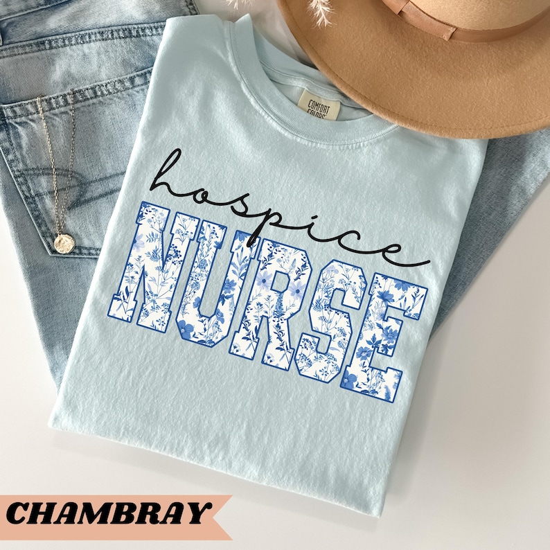May include: Light blue t-shirt with the text "hospice NURSE" in white letters with a blue floral pattern. The shirt is made of a soft, comfortable fabric.